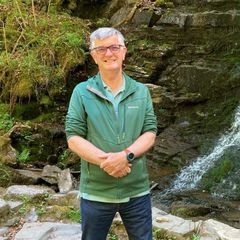 Andrew Date, smiling, standing in front of a waterfall in a green fleece