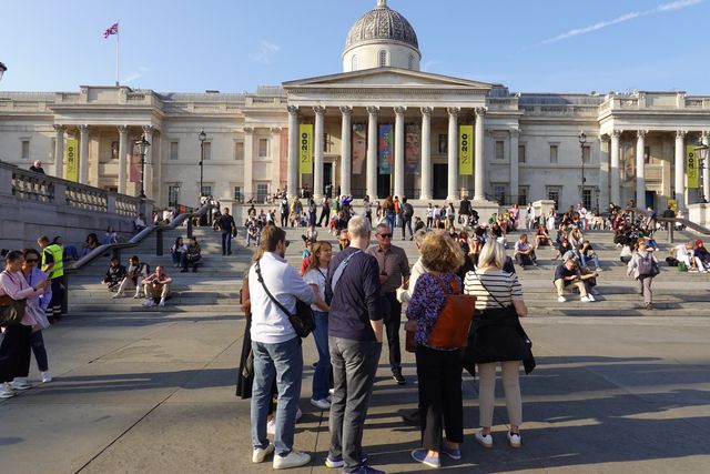 A walking group gathers in Trafalgar Square in late afternoon sun, with the National Gallery behind