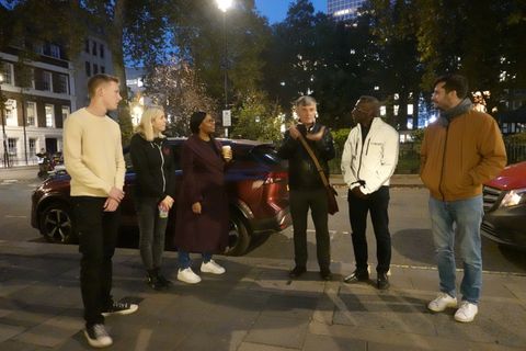 A walking group listens to their guide on a street corner by Soho Square in the evening