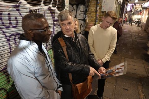 A guide shows walkers an archive photograph beside a graffiti-covered shutter in a Soho alley