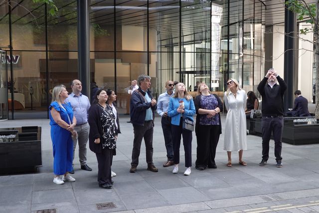 A group of colleagues on a wellbeing walk stop outside a glass office building in the City of London to look up at the architecture