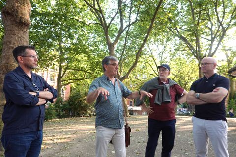 A guide gestures as he tells a story to a team of colleagues under the trees of a leafy East London square